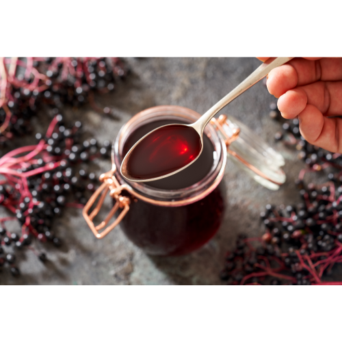 hand holding a spoonful of elderberry syrup over an open mason jar. Bunches of fresh elderberries are surrounding the jar giving a medicinal and herbal DIY aesthetic.